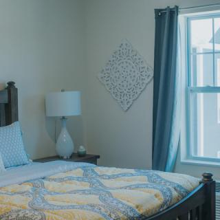 A cozy bedroom with a neatly made bed, accented by a lamp and decorative wall piece near the window.