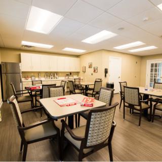 A well-lit dining area with multiple tables and chairs, featuring a kitchenette in the background.
