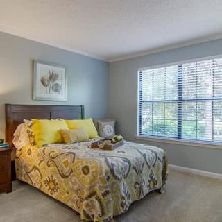 A cozy bedroom with a neatly made bed featuring yellow accents, a large window with blinds, and tasteful decor.