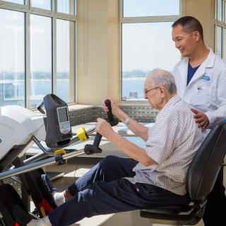 An elderly man receives guidance from a healthcare professional while using exercise equipment in a rehabilitation setting.