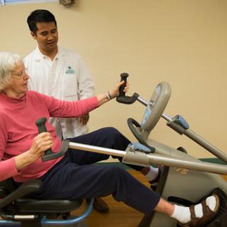An elderly woman engages in a physical therapy session with the assistance of a healthcare professional, using specialized exercise equipment.