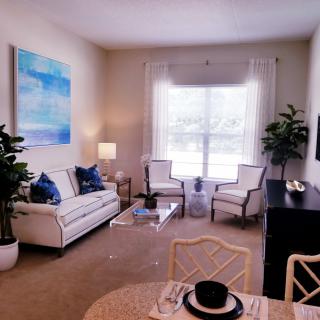 A cozy and elegantly furnished living room with a large window, featuring a sofa, chairs, artwork, and decorative plants.