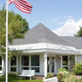 A quaint white house with an American flag in the front garden, accompanied by trees and a bench under a clear blue sky.