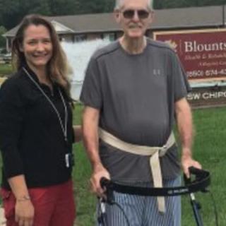 An elderly man using a walker stands outdoors next to a woman, with a building and sign in the background.