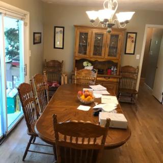 A dining room with a large wooden table surrounded by chairs, papers scattered on the table, and a fruit bowl in the center. A china cabinet is against the wall, and a chandelier hangs above.