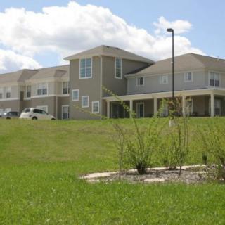 A modern residential building with a grassy foreground and parked cars under a cloudy sky.