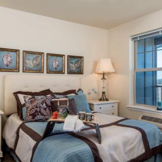 A cozy bedroom with a neatly made bed, decorative pillows, a bedside lamp, and framed artwork above the headboard.