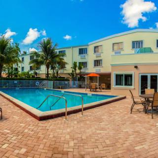 An outdoor swimming pool area at a hotel, surrounded by palm trees and patio furniture on a sunny day.