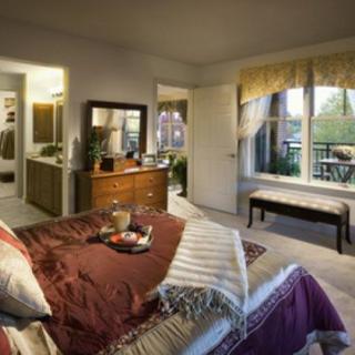 A cozy bedroom with warm decor and a comfortable bed, featuring a dresser, mirror, and large windows with a view of the balcony.
