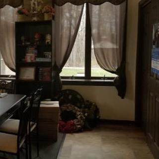 Cozy living room with a shelf, books, and large windows draped with curtains.