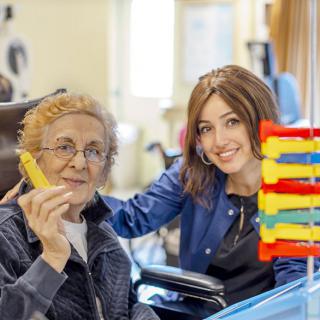 A caregiver interacting with an elderly person, both smiling in a nursing home setting.