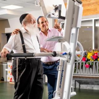 A senior man receiving assistance with a rehabilitation exercise machine in a healthcare facility.