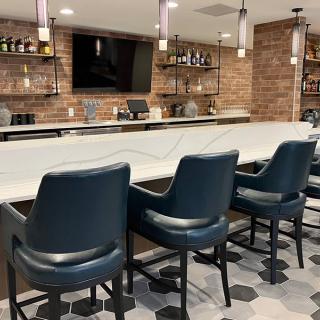 A modern bar with high chairs and a sleek countertop, featuring shelves of drinks and a TV on the wall.