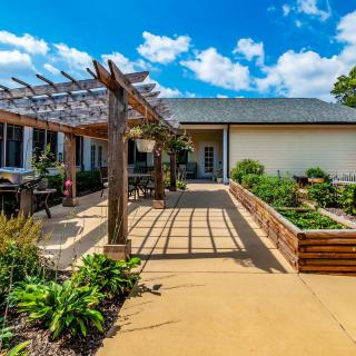 A sunny outdoor patio with a pergola, surrounded by lush greenery and raised flower beds.