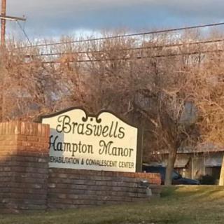 A sign for Braswells Hampton Manor in front of a brick wall, with trees and a building in the background.