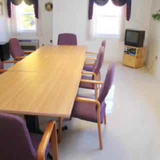 A conference room with a long meeting table and chairs, featuring a TV on a stand and natural light coming through the windows.