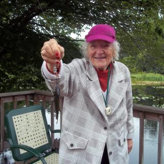 An elderly person smiles while holding a small fish on a deck by the water.