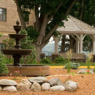 A peaceful garden setting featuring a three-tiered fountain and a wooden gazebo surrounded by greenery.