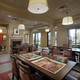 A cozy dining area with tables and chairs arranged under ceiling lights, featuring large windows that allow natural light to illuminate the space.