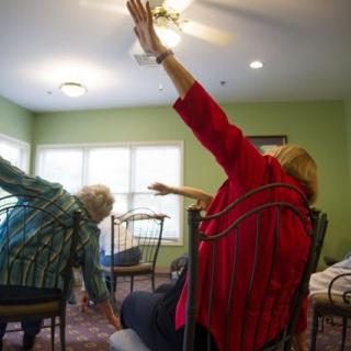 Seniors participating in a seated exercise class, reaching their arms to the ceiling.