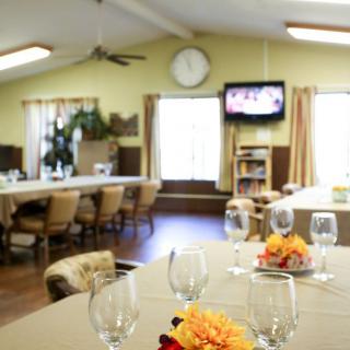 A dining room with tables set for an event, featuring elegant glassware and floral centerpieces, with a television in the background.