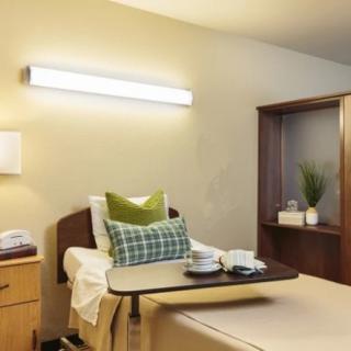 A clean and well-organized hospital room featuring a bed with green pillows, a wooden cabinet, and bright lighting.