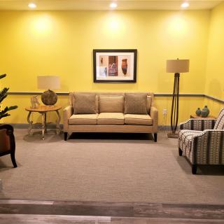 A cozy living room with modern furniture, featuring a sofa and armchair against a bright yellow wall adorned with artwork.