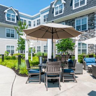 An outdoor patio area with a table and chairs under an umbrella, set in front of a residential building on a sunny day.