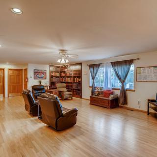 A spacious living room with hardwood flooring, featuring cozy recliners, built-in bookshelves, and large windows.