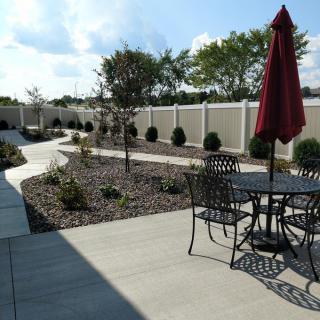 An outdoor patio with a table, chairs, and an umbrella, surrounded by landscaped areas and a pathway.