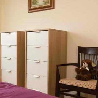 A cozy bedroom featuring a pair of dressers, a chair with a teddy bear, and a painting on the wall.
