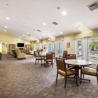 A spacious and well-lit living area with multiple tables and chairs, featuring large windows and a comfortable atmosphere.