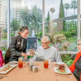 A waitress serves food to two elderly patrons at a restaurant with a garden view.