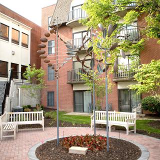 A tranquil courtyard featuring a central sculpture, surrounded by benches and greenery, adjacent to a brick building.