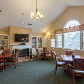 A cozy living room with multiple tables and chairs, a television above a fireplace, bookshelves on either side, and large windows providing natural light.