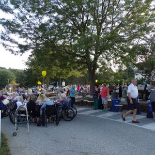 A lively community gathering taking place outdoors, with people enjoying a summer evening under the trees.