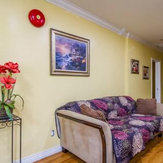A cozy living room with a floral-patterned couch, vibrant red flowers on a stand, and artwork on the yellow walls.