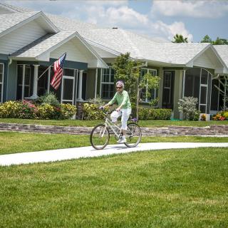 A person rides a bicycle along a sidewalk in a suburban neighborhood on a sunny day, with houses and an American flag in the background.
