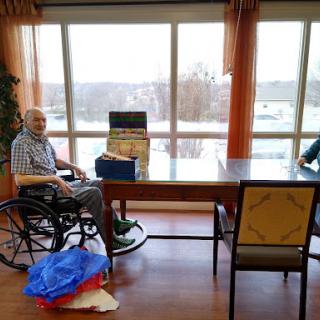 An elderly man sitting in a wheelchair beside a table with a view out the window.