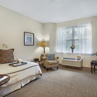 A cozy hotel bedroom with a neatly made bed, armchair, and sunlight streaming through the window.