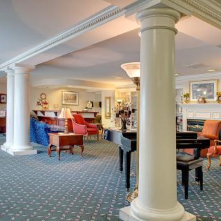 A spacious lounge area with elegant columns, a piano, and colorful chairs arranged on a patterned carpet.