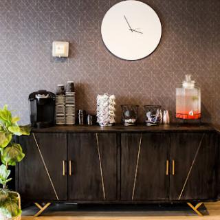 A stylish coffee station featuring a modern black cabinet, wall clock, and a drinks dispenser, complemented by decorative plants.