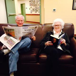 An elderly couple sitting on a couch, each engrossed in reading; one with a newspaper and the other with a book.