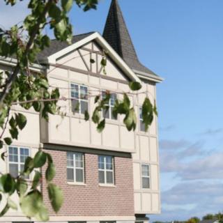 A building with a pointed roof and several windows, partially obscured by green leaves against a blue sky.