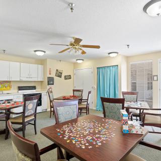 A cozy dining area with tables set up for games and meals, featuring a ceiling fan and a small kitchen area.