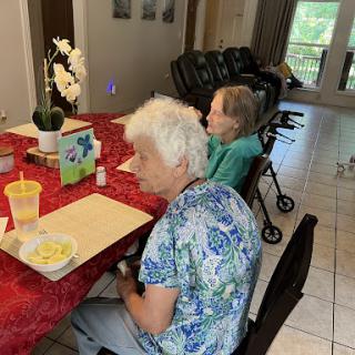Two elderly women seated at a dining table in a care facility, engaged in conversation.