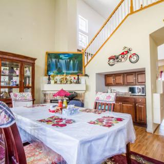 A well-decorated dining room featuring a table set with a white cloth, surrounded by wooden chairs. The space includes a cabinet, wall paintings, and a decorative motorcycle on the wall near the staircase.