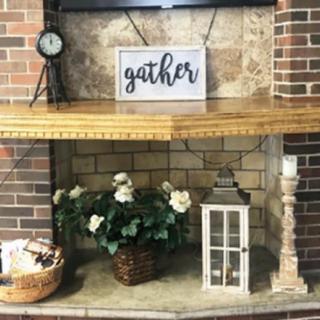A cozy fireplace adorned with decorative items, including a sign that says 'gather', a vase of flowers, and a lantern.