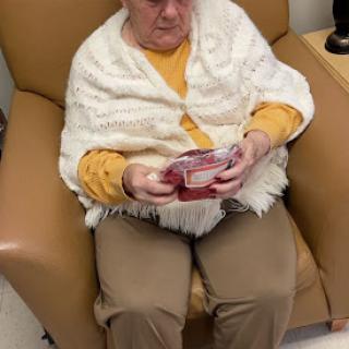 An elderly woman sits comfortably in a chair, engrossed in reading something while wrapped in a cozy blanket.