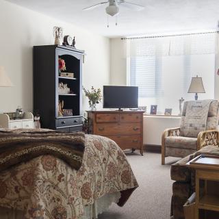 A cozy bedroom with classic decor, featuring a bed, armchair, television, and various furniture pieces.